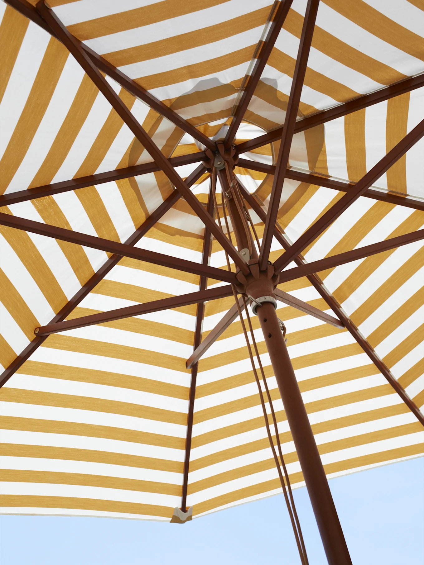 Undersidan av ett gul- och vitrandigt parasoll, som visar trästommen mot en blå himmel.