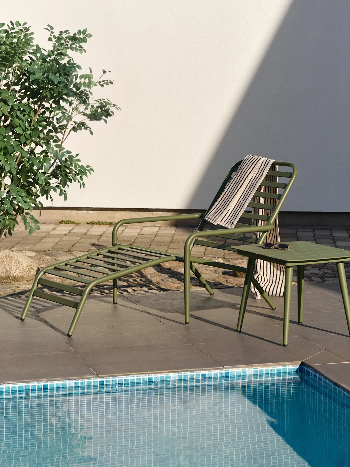 Grön solsäng och sidobord vid en pool med en randig handduk och solglasögon.
