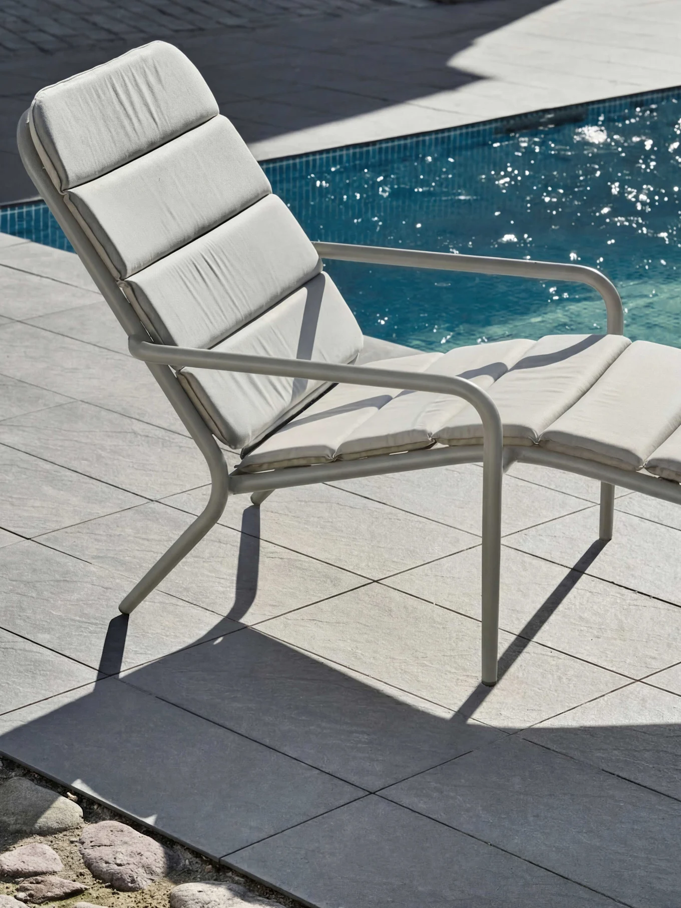 Ljusgrå utomhus solsäng med dynor vid en pool, på grå plattor med solsken.