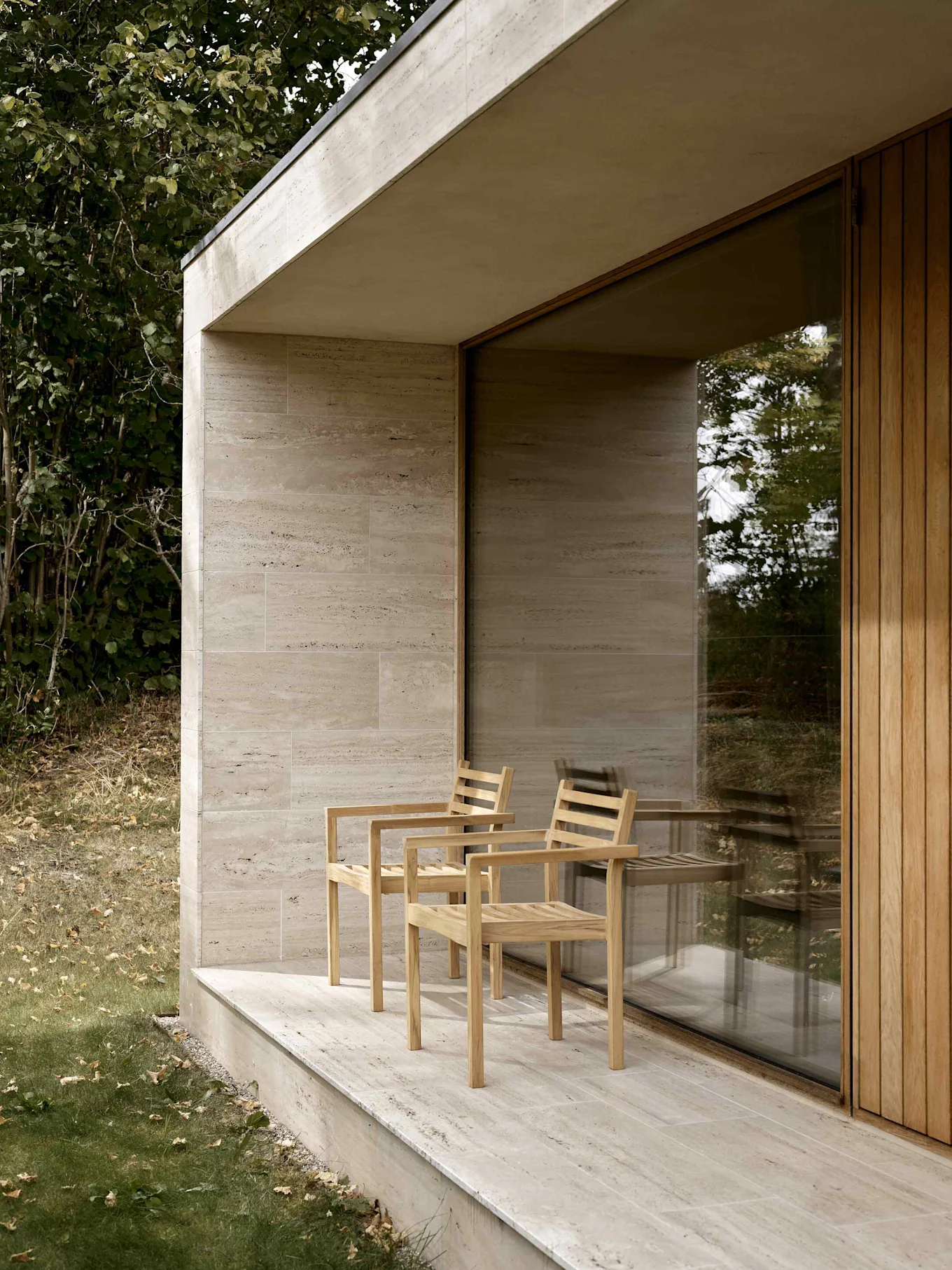 Två ljusa träläktstolar på en beige stenuteplats vid en modern byggnad med glasfönster och träpanel.