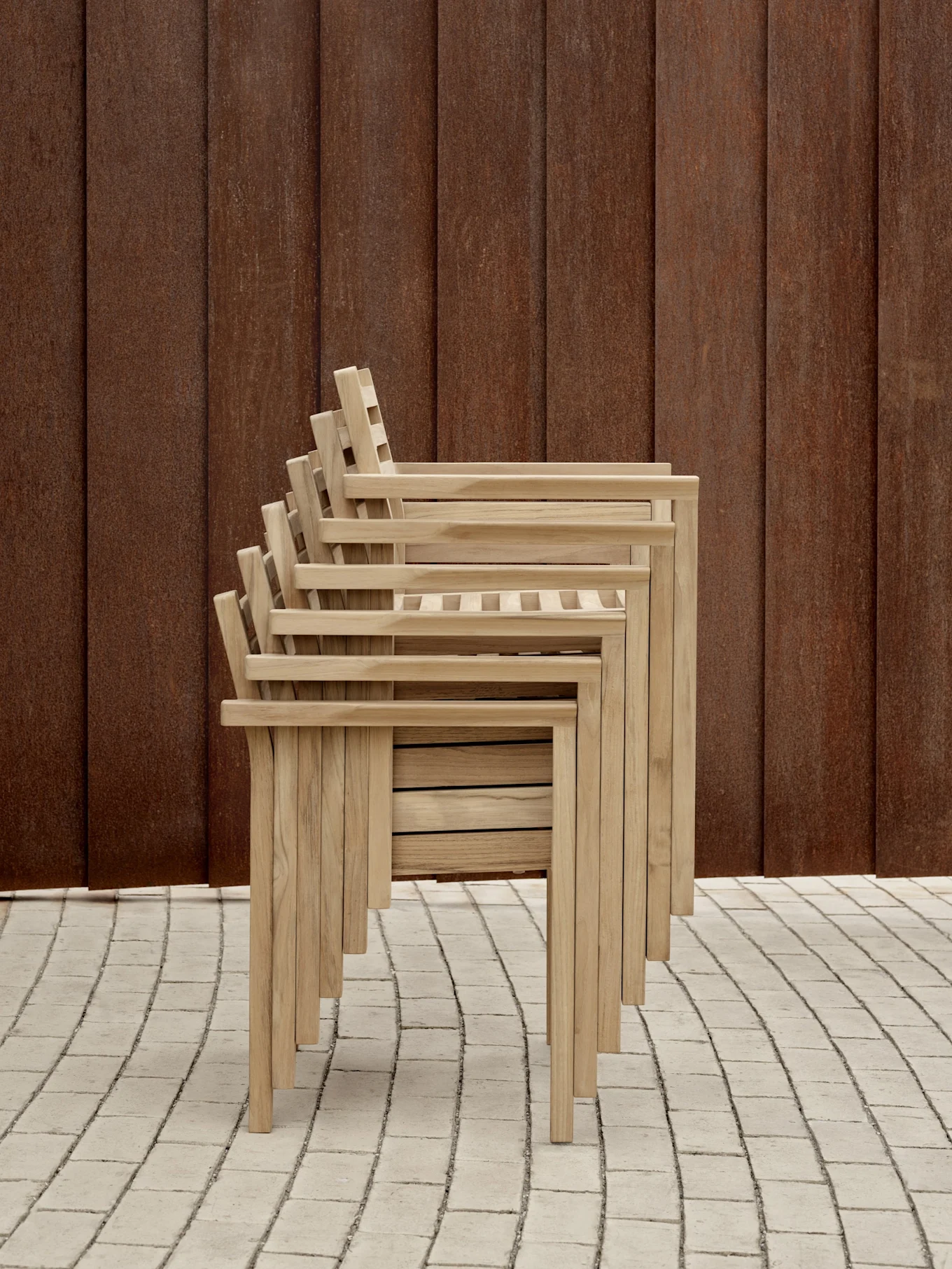 En stapel med ljusa trädgårdsstolar med ribbad rygg och sits, framför en rostbrun vägg på ett klinkerlagt golv.