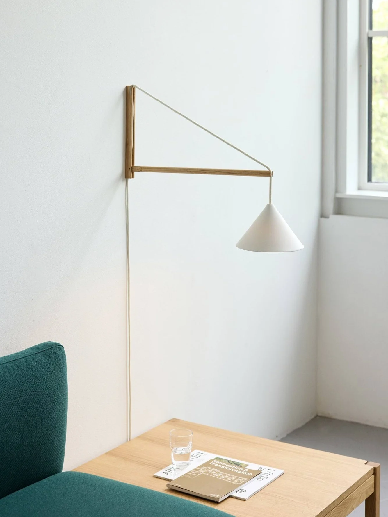 Väggmonterad lampa med vit konformad skärm och träarm ovanför ett soffbord med glas och tidningar, bredvid en grön soffa.