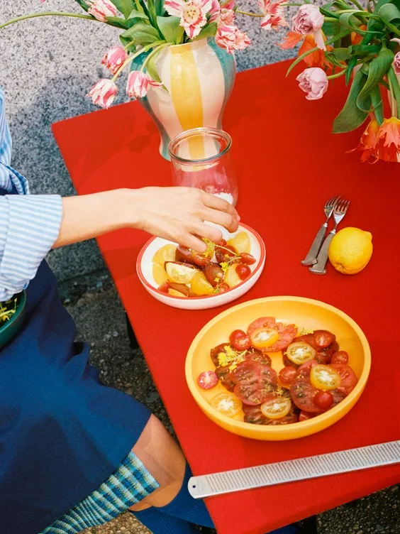 En persons hand sträcker sig efter skivad frukt på ett livfullt rött bord med en färgglad tomatsallad och tulpaner.
