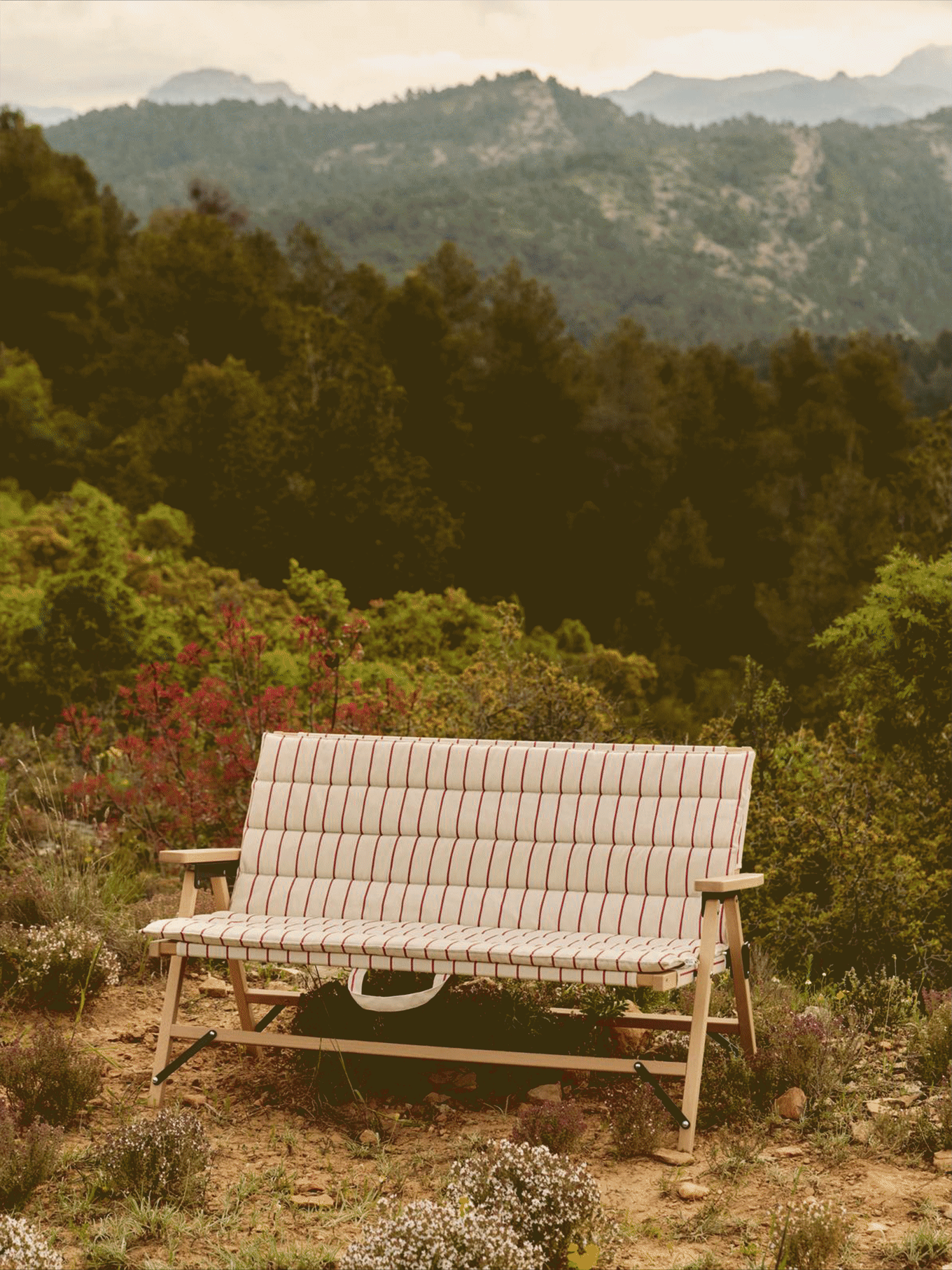 Randig utomhusbänk med ljus träram i ett bergslandskap med träd.