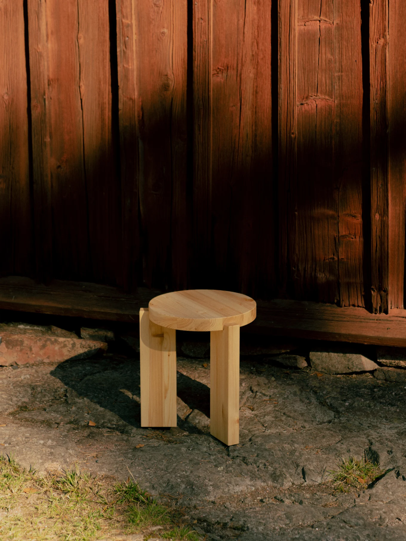 Liten, ljus träpall med rund topp och tjocka ben på en sten uteplats framför en röd trävägg.