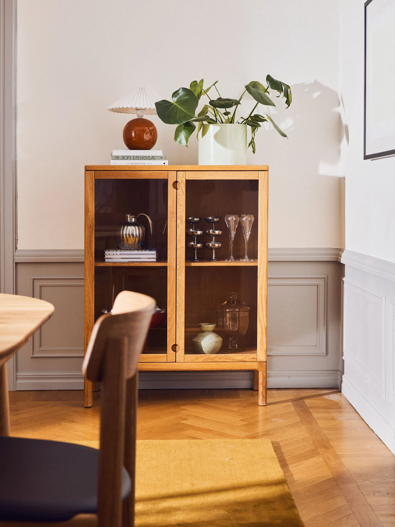Vitrinskåp i ek med glasdörrar, inredningsdetaljer, en lampa, böcker och en växt. Rummet har trägolv och en gul matta.