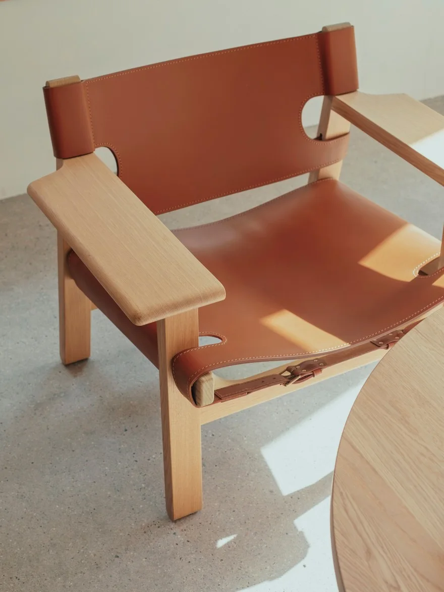 Stol med ljus träram och karamellbrun lädersits och rygg, med synliga spännen. Ett runt träbord är delvis synligt.