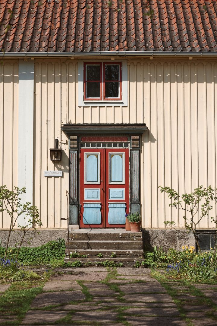 Bild som visar den byggnad Siv och Carl Malmsten bodde i när Capellagården en gång startades 1960. En stor träbyggnad i gult med vackert målad dörr i rött och ljusblått.