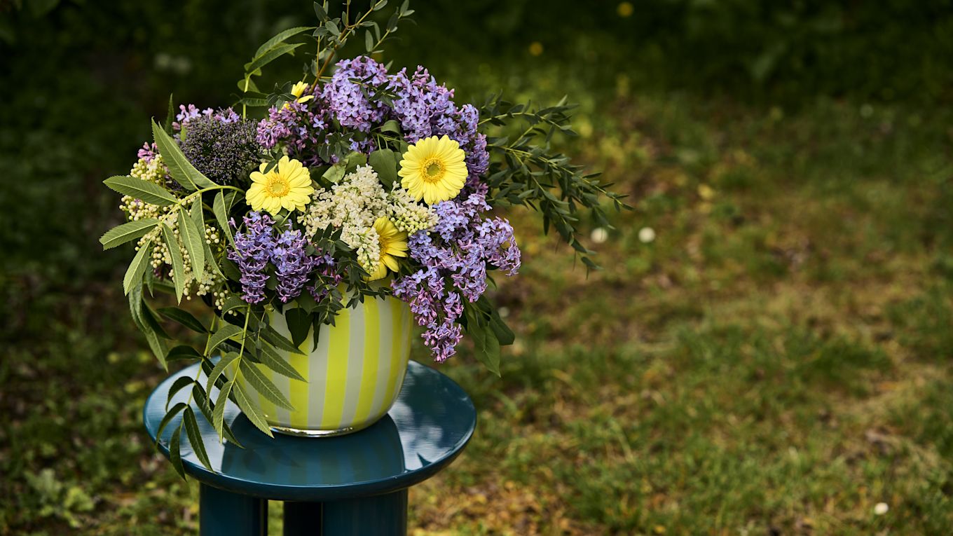 Bild som visar en blombukett i Juice Wide vas som är gul- och vitrandig i glas från varumärket HAY, placerad på JA Tung sidobord i blått från &Tradition.