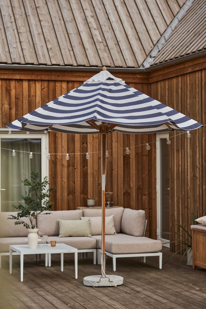 Blåvitt randigt parasoll från varumärket 1898, här på ett stilrent trädäck där det också står en loungesoffa i vitt och beigt.