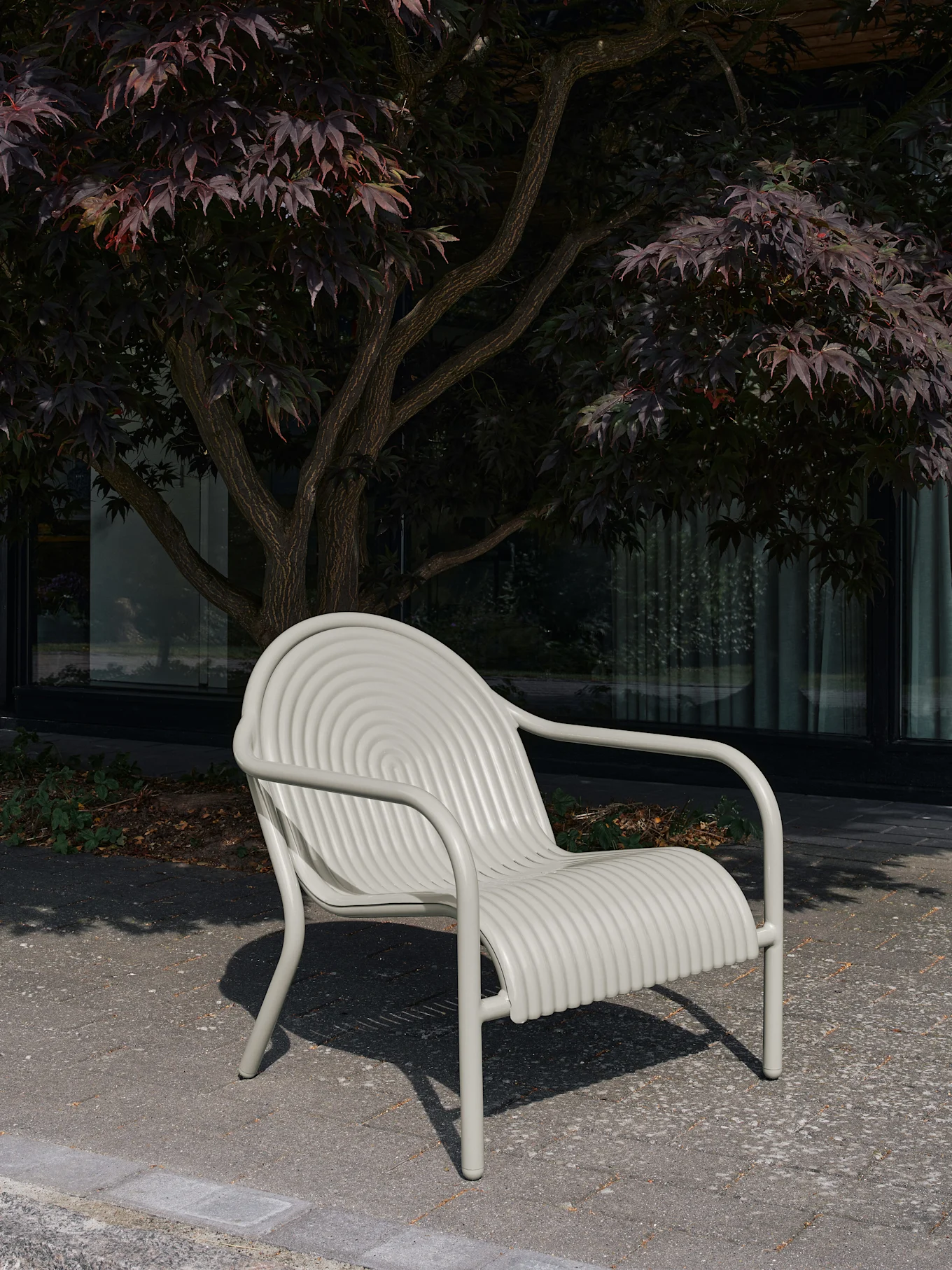 En ljusgrå utomhusstol med ryggstöd med koncentriska cirklar och sits med horisontella räfflor, placerad på plattor med ett träd bakom.