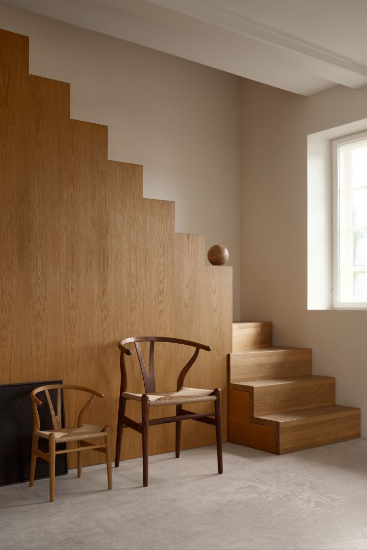 Helbild på de populära designstolarna CH24 originalstol & barnstol bredvid varandra i hallmiljö med trappa.