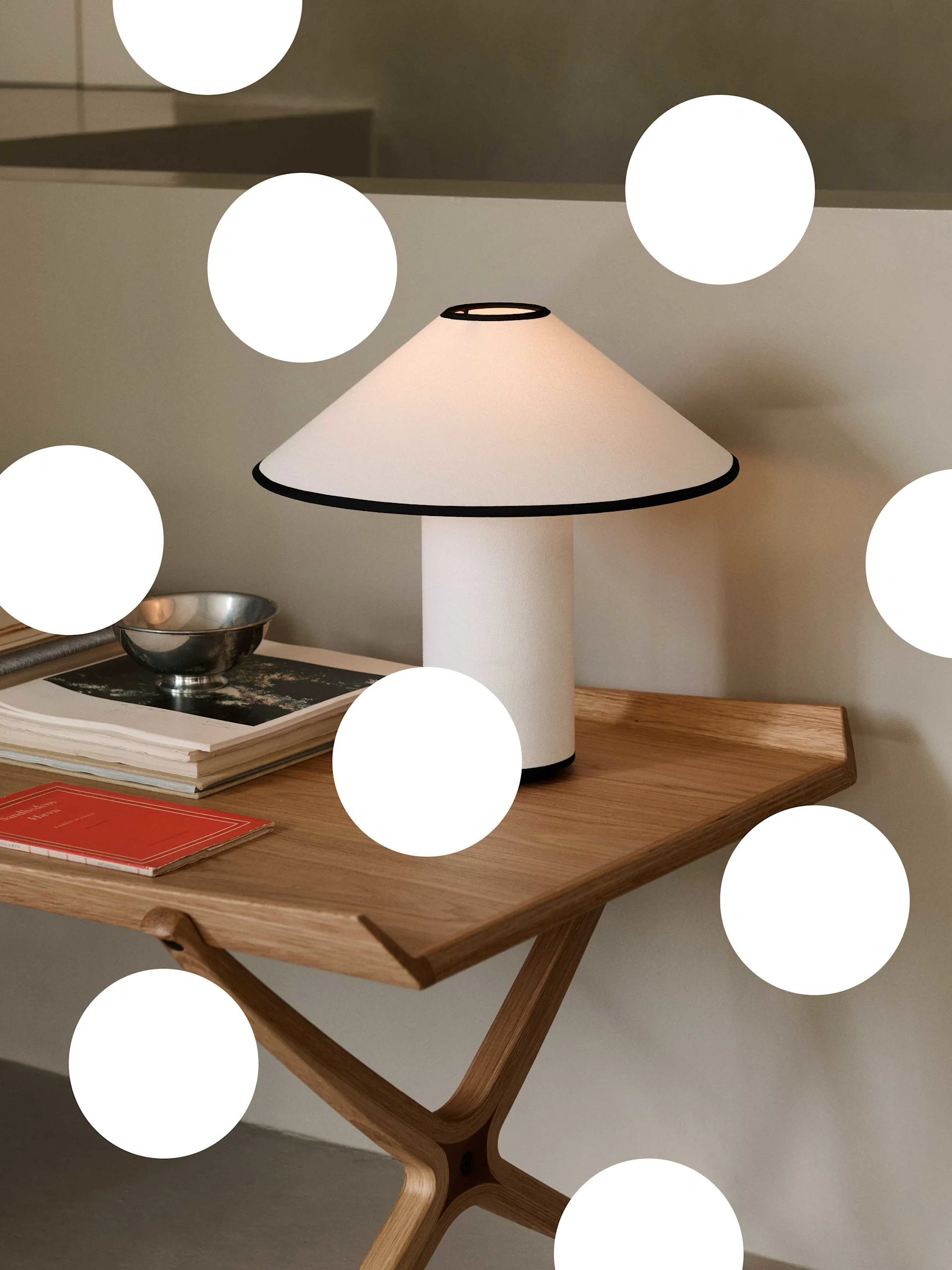 En modern vit bordslampa med svart kant på ett ljust träbord, bredvid böcker och en silver skål.