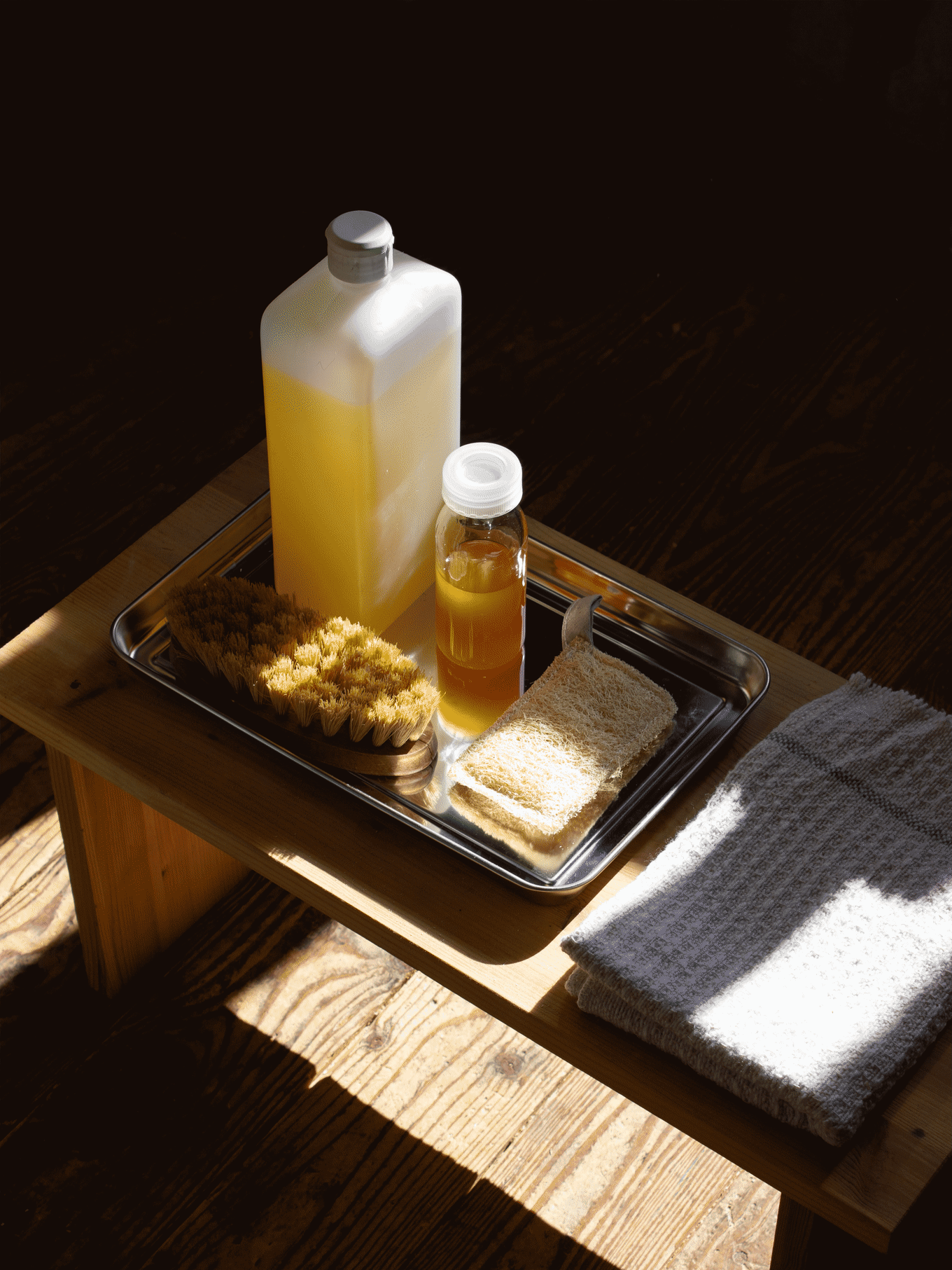 Badartiklar på silverbricka på träpall: gul flaska, liten bärnstensfärgad flaska, borste, luffa, handduk.