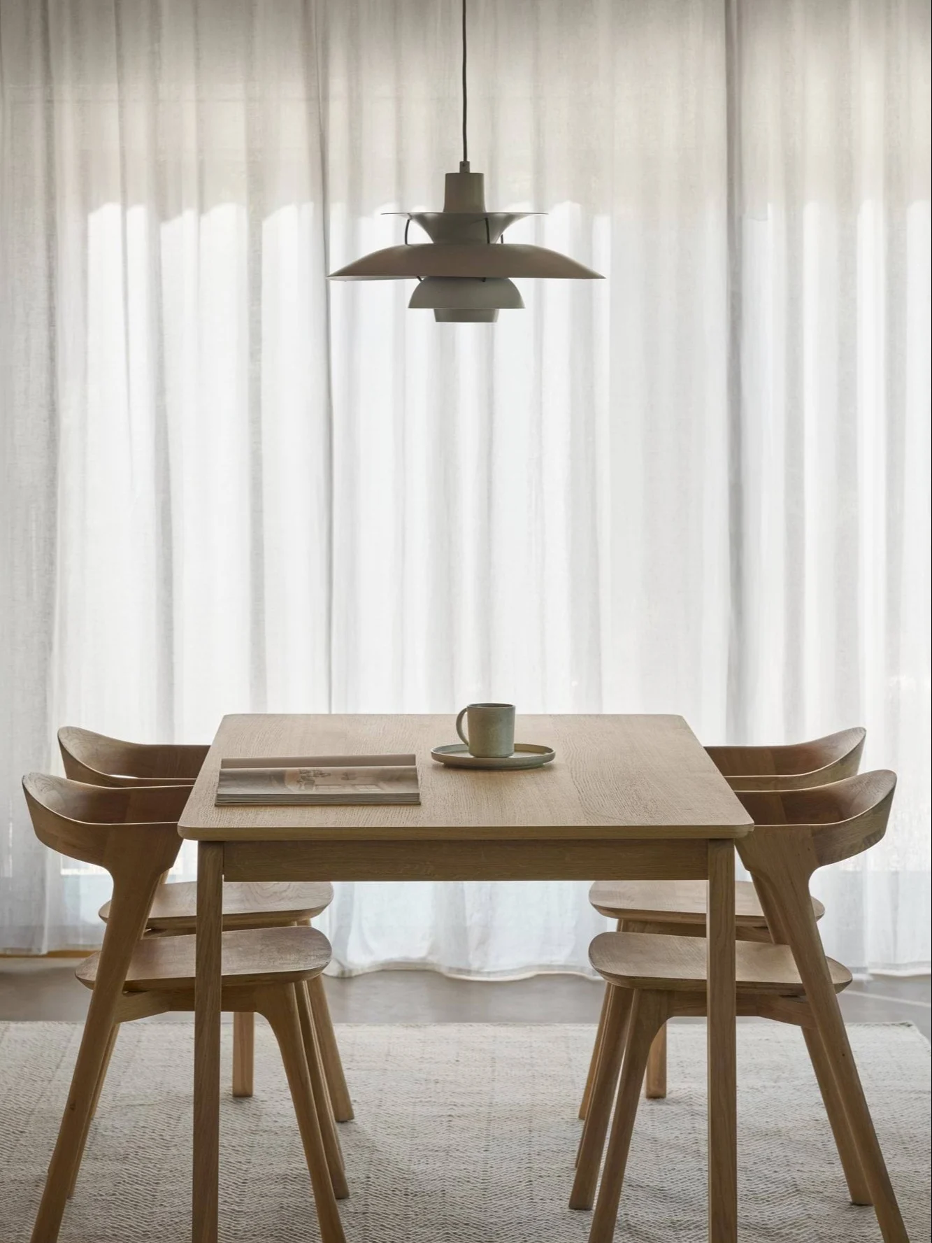 En matplats med ett ljust träbord, fyra matchande stolar och en unik taklampa framför vita gardiner.