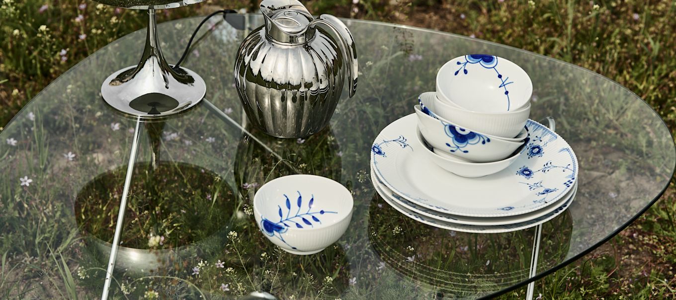 Bild som visar blåvitt porslin från Royal Copenhagen, serien blue elements placerade på Pedrera soffbord i glas från Gubi, bredvid står Bernadotte termoskanna och Panthella portabel lampa i krom från Louis Poulsen, allt placerat på en blommande sommaräng.